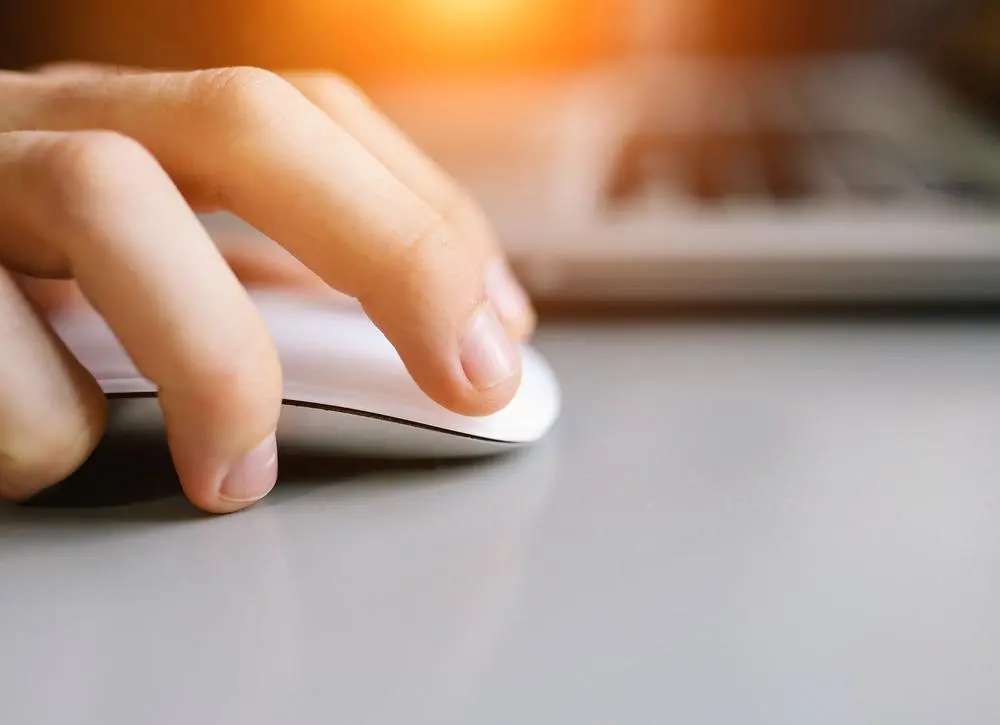 A close-up of a hand using a white computer mouse on a gray surface, with part of a laptop keyboard visible in the background and a warm light shining from the top.