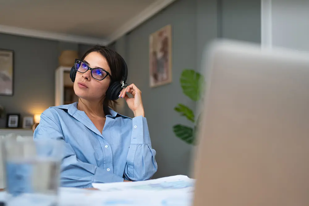 A woman wearing glasses and headphones sits at a desk, looking thoughtful. She is in a home office setting with a laptop, papers, and a plant visible in the background.