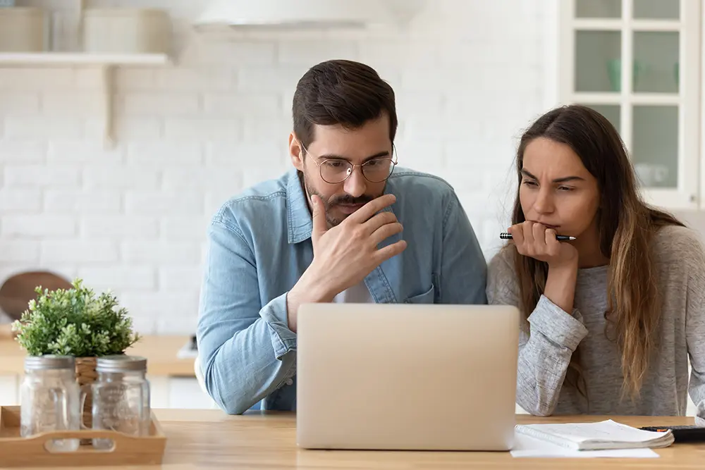 A man and a woman sit at a table in a kitchen, looking thoughtfully at a laptop screen. The woman holds a pen, and papers are spread out in front of them. A small plant is on the table.