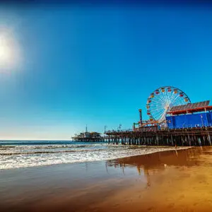 A sunny day at the beach with gentle waves, a wooden pier extending over the ocean, and a colorful Ferris wheel and buildings on the pier under a clear blue sky.
