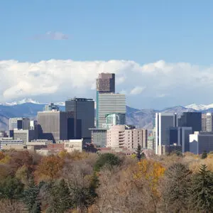 Downtown city skyline with modern skyscrapers, trees with autumn foliage in the foreground, and snow-capped mountains in the background under a blue sky with clouds.