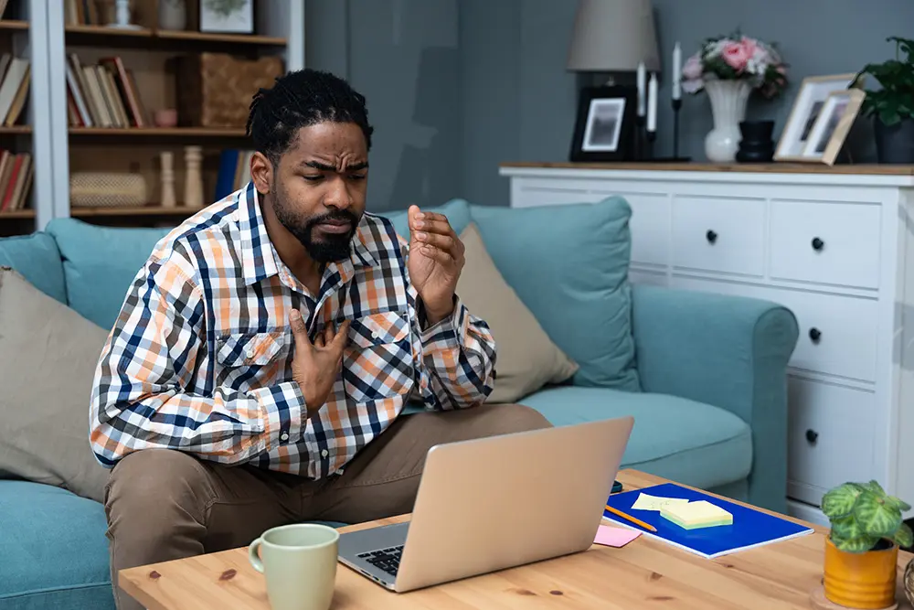 A man sitting on a couch holds his chest and looks distressed while staring at a laptop on a coffee table, appearing to experience discomfort or pain. The setting is a cozy living room with bookshelves and home decor.