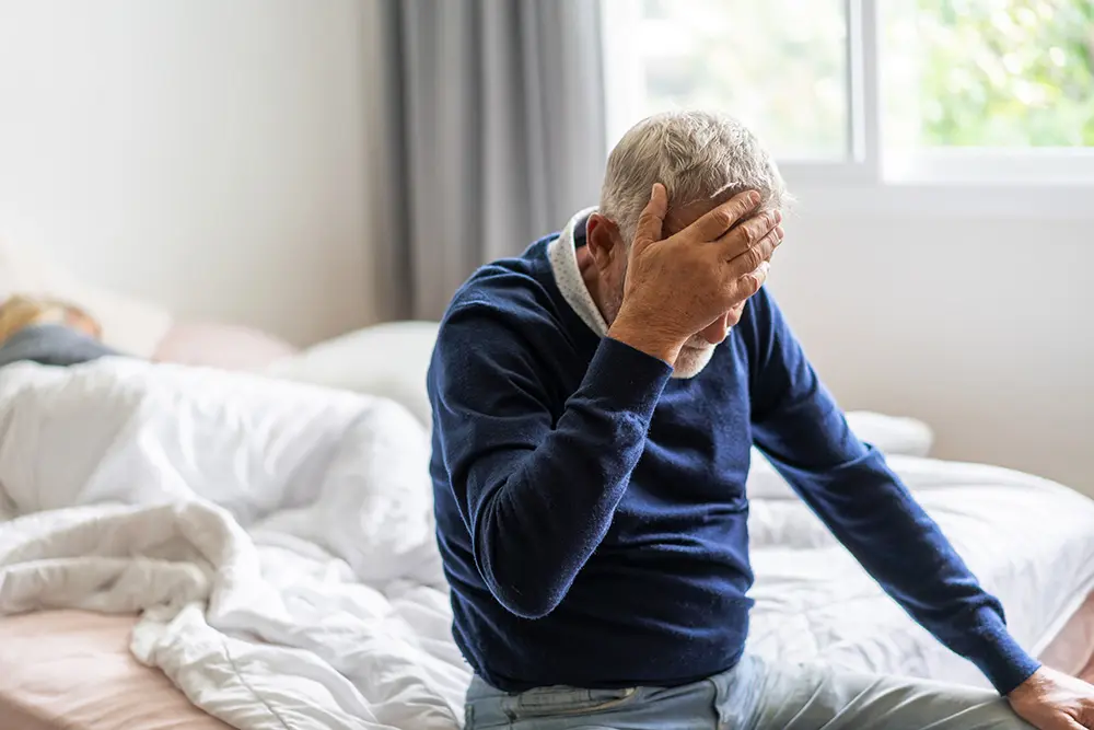 An older man sits on the edge of a bed with his hand on his forehead, looking distressed. A person lies under the covers in the background. The room is softly lit with natural light from a window.