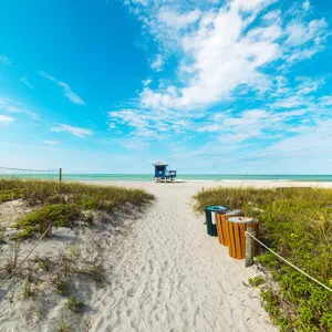 A sandy path leads to a lifeguard tower on a beach, bordered by grass and bushes, with blue sky and scattered clouds overhead. Trash and recycling bins are placed along the path.