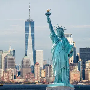 The Statue of Liberty stands in the foreground with the New York City skyline and One World Trade Center visible in the background under a clear sky.