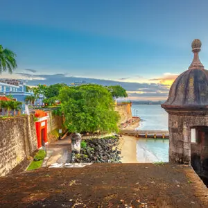 View of historic city walls, a sentry box, and the red San Juan Gate overlooking a small beach and the ocean under a colorful sky in Old San Juan, Puerto Rico.