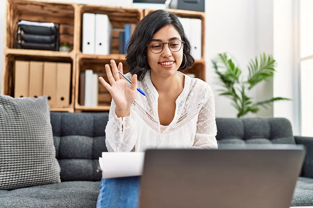 A woman with glasses smiles and waves at her laptop while sitting on a sofa, holding a pen and notebook, in a bright living room with shelves and a plant in the background.