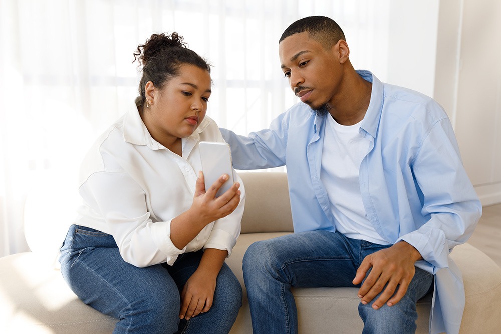 A woman and a man sit close together on a couch, looking seriously at a smartphone the woman is holding. The man has his arm gently around her shoulders. Both appear concerned or thoughtful.